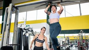 Two women practicing pull-ups in a modern indoor gym environment, emphasizing fitness and training.